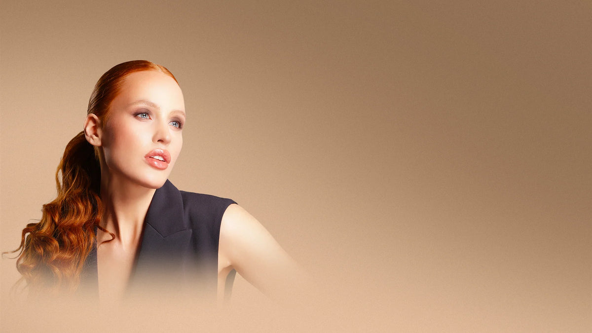 Woman with copper-red hair tied back wearing a black sleeveless blazer against a warm brown background.